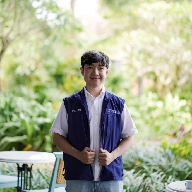 Rahmat Fajri standing in a garden, wearing a navy vest over a white shirt